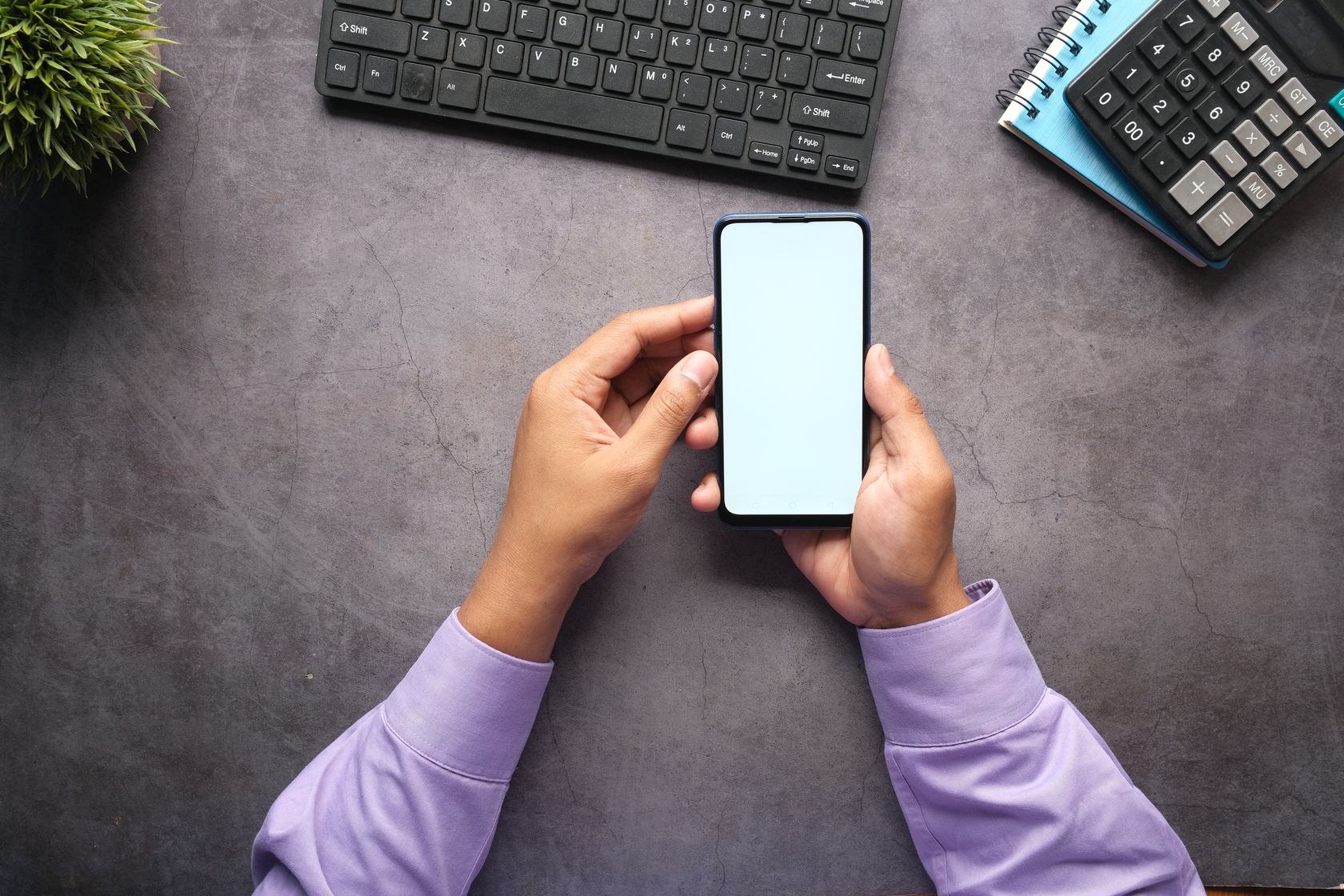 Dłonie trzymające smartfon z pustym ekranem obok klawiatury laptopa i kalkulatora na biurku, symbol pracy nad aplikacją mobilną i automatyzacją procesów w firmie